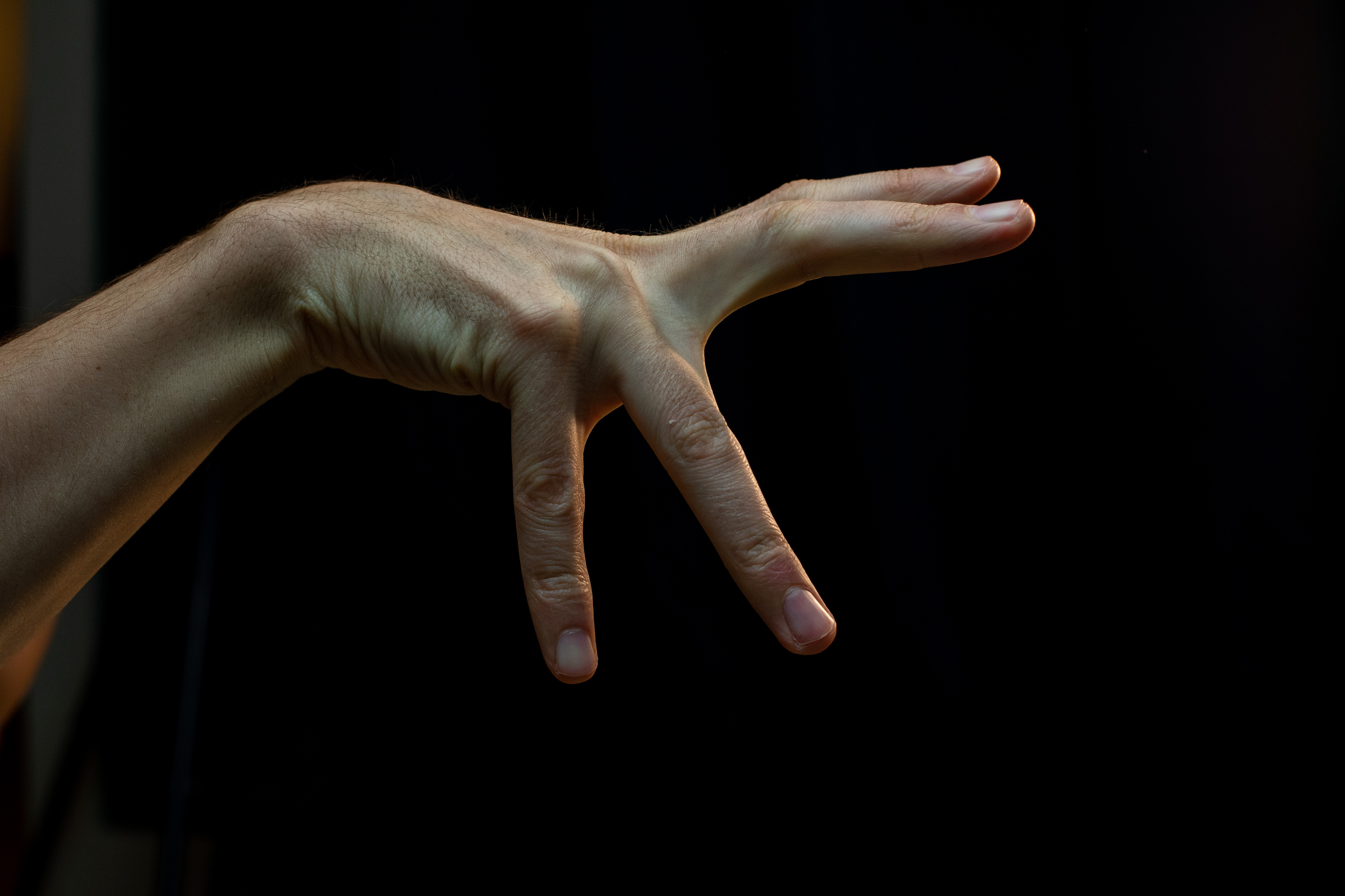 a hand is extended against a black background, illuminated with artificial light. the hand is positioned with the palm facing the viewer, and the fingers are extended and slightly spread apart. the first three fingers are fully extended, while the pinky finger is curled inward. the image has a studiolike quality, with a focus on the hand and its details.