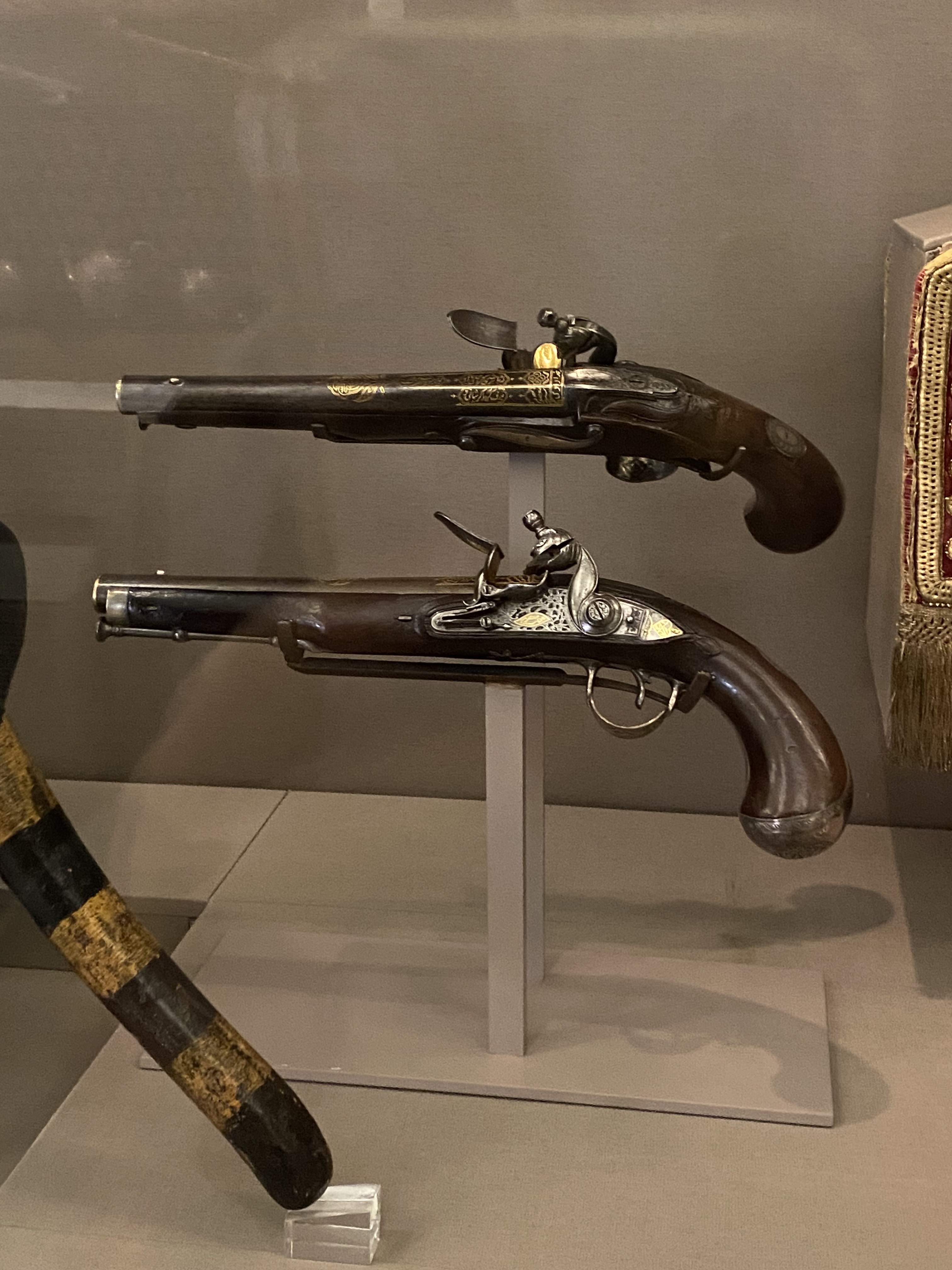 the image shows two flintlock pistols on display in a museum. the pistols are resting on individual stands within a glass case. both pistols feature ornate metalwork and dark wooden stocks, showcasing their antique and decorative nature. a cane is partially visible in the corner.