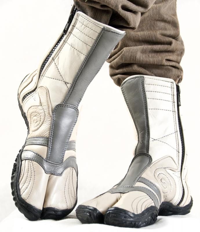 a closeup shot of a person wearing a pair of beige and white leather tabi boots with grey accents. the boots have a splittoe design and zipper closures. the person is also wearing brown pants. the background is a plain white surface.