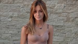 a closeup portrait of a young, attractive blonde woman standing in front of a stone wall. she's wearing a light brown tank top and a delicate necklace. her expression is pleasant and neutral. the lighting suggests it might be outdoors on a sunny day.  the overall feel is casual and confident.