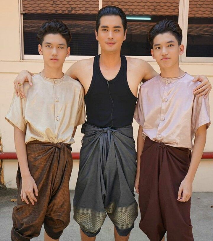 three young men are standing together outdoors, smiling. the man in the middle is wearing a black tank top and grey pants. the men on either side of him are wearing traditional thai clothing: a white shirt and brown pants or a brown shirt and brown pants.  the man in the middle has his arms around the other two men.  they are all smiling and looking happy.
