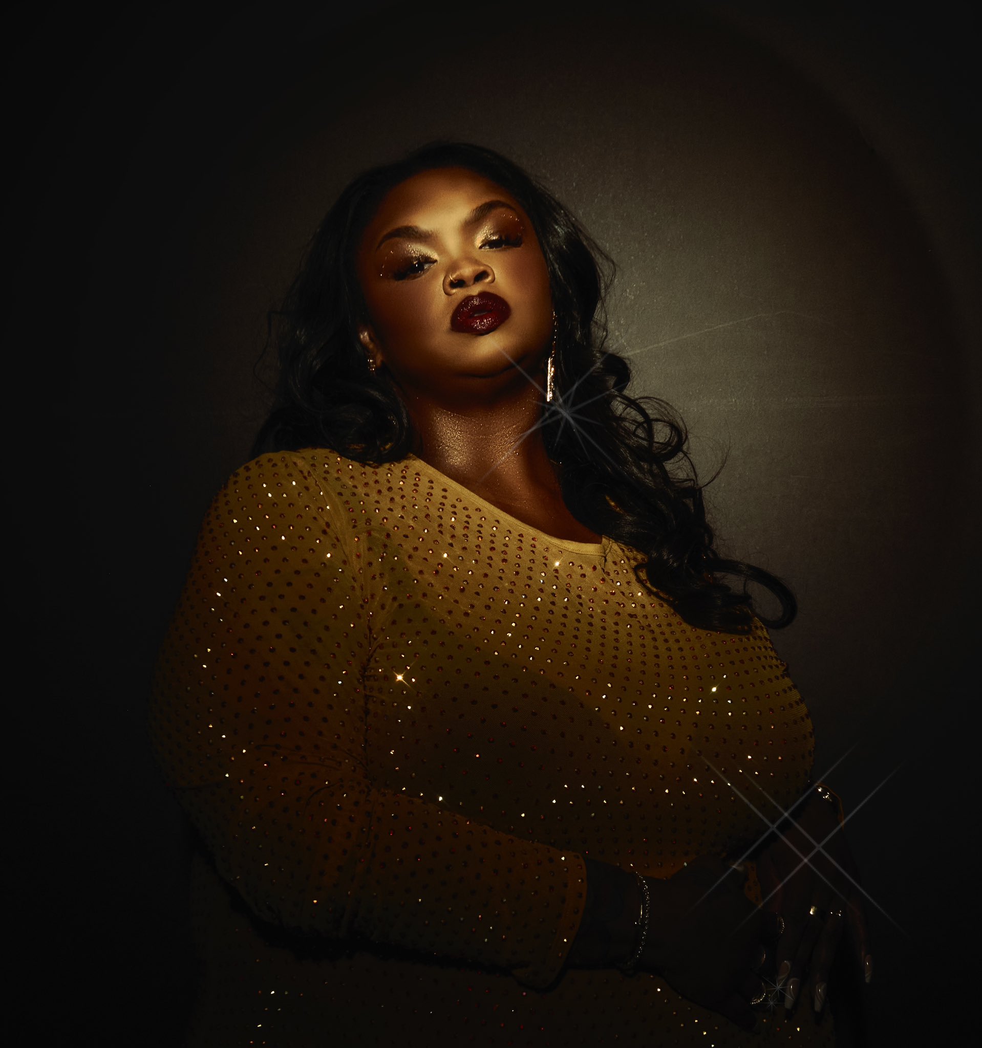 a young woman in a gold sequin dress stands in a darkened studio. she looks directly at the camera with a serious expression. her makeup is dramatic and highlights her beautiful features. her hair is worn long and flowing.  the image is lit from the front, creating a dramatic effect.  the woman is likely a model or actress, as her pose and expression are confident and striking. the gold sequins on her dress sparkle and shimmer in the light, creating a glamorous look.