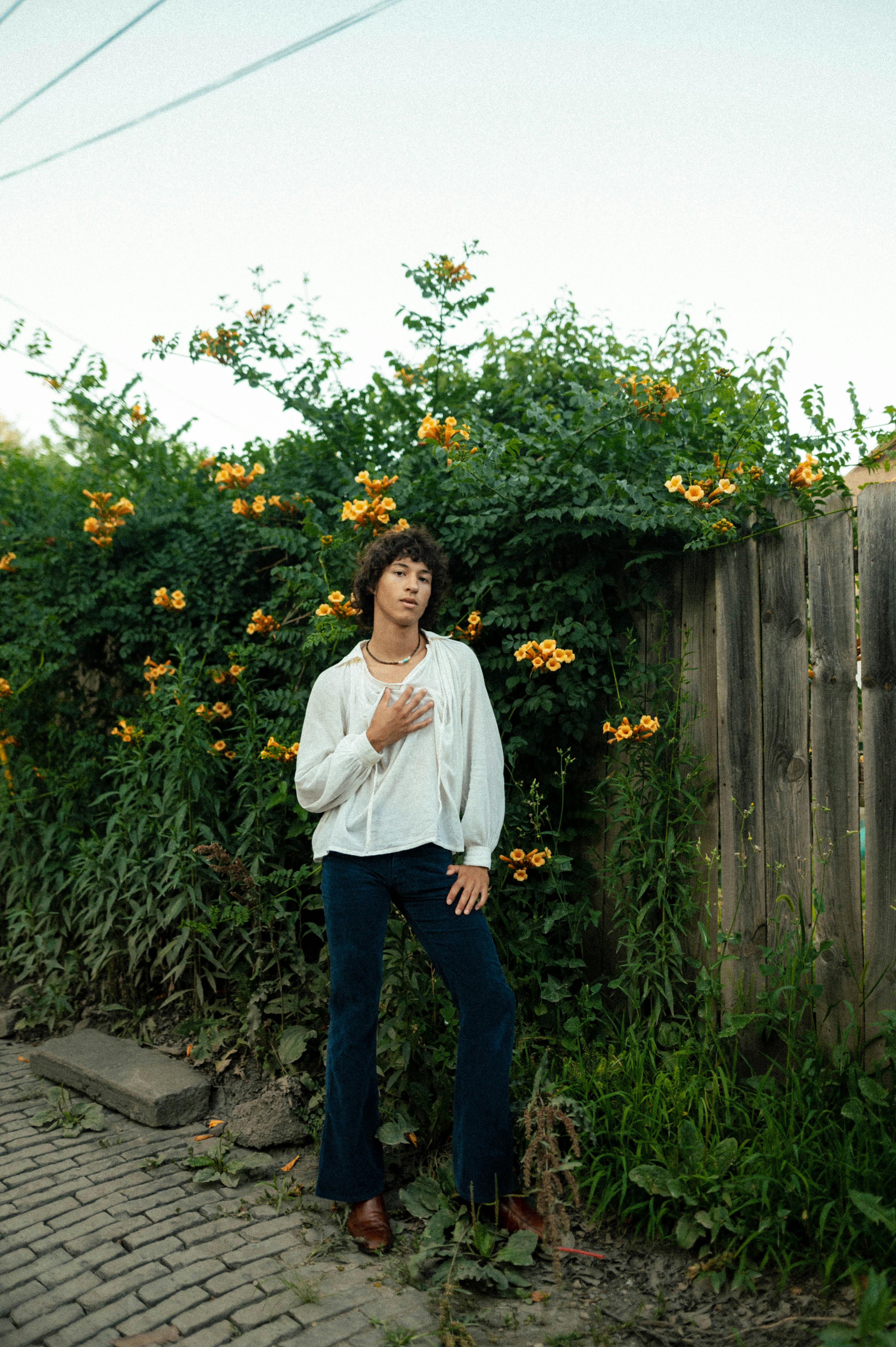 a young man stands in a relaxed pose in a narrow alleyway. he's wearing a white, loosefitting shirt and dark teal bellbottom pants, creating a 70s retro style. his dark curly hair and calm expression add to the vintage vibe. he stands against a backdrop of a wooden fence covered with a lush green vine bearing bright yellow flowers. the brick pavement adds texture to the scene. the overall feel is calm and slightly nostalgic.