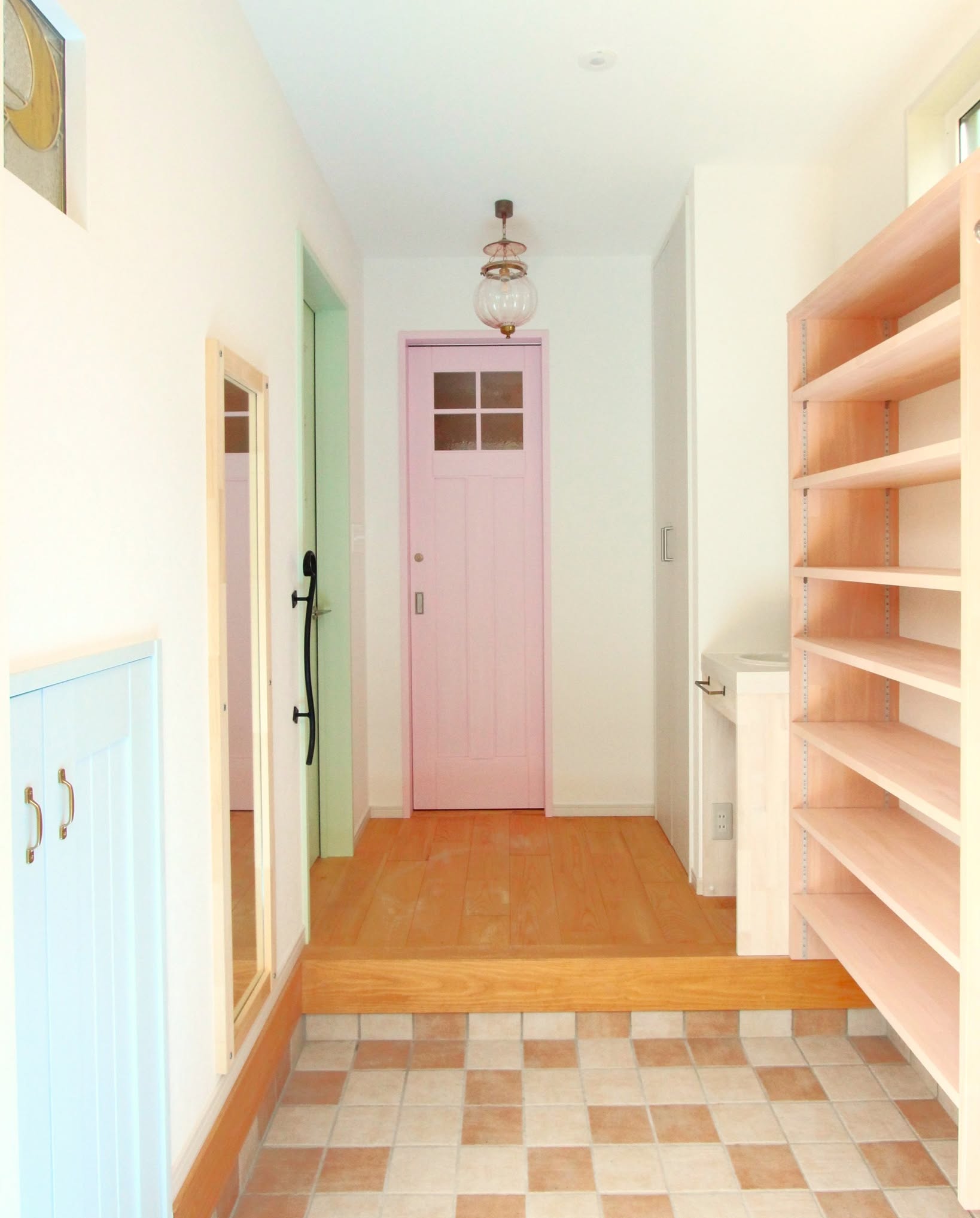 an interior shot shows a hallway with a pink door at the end, a light fixture hanging from the ceiling, a mirror, blue cabinets on the left, shelving on the right, and a checkered tile floor with a wooden platform.