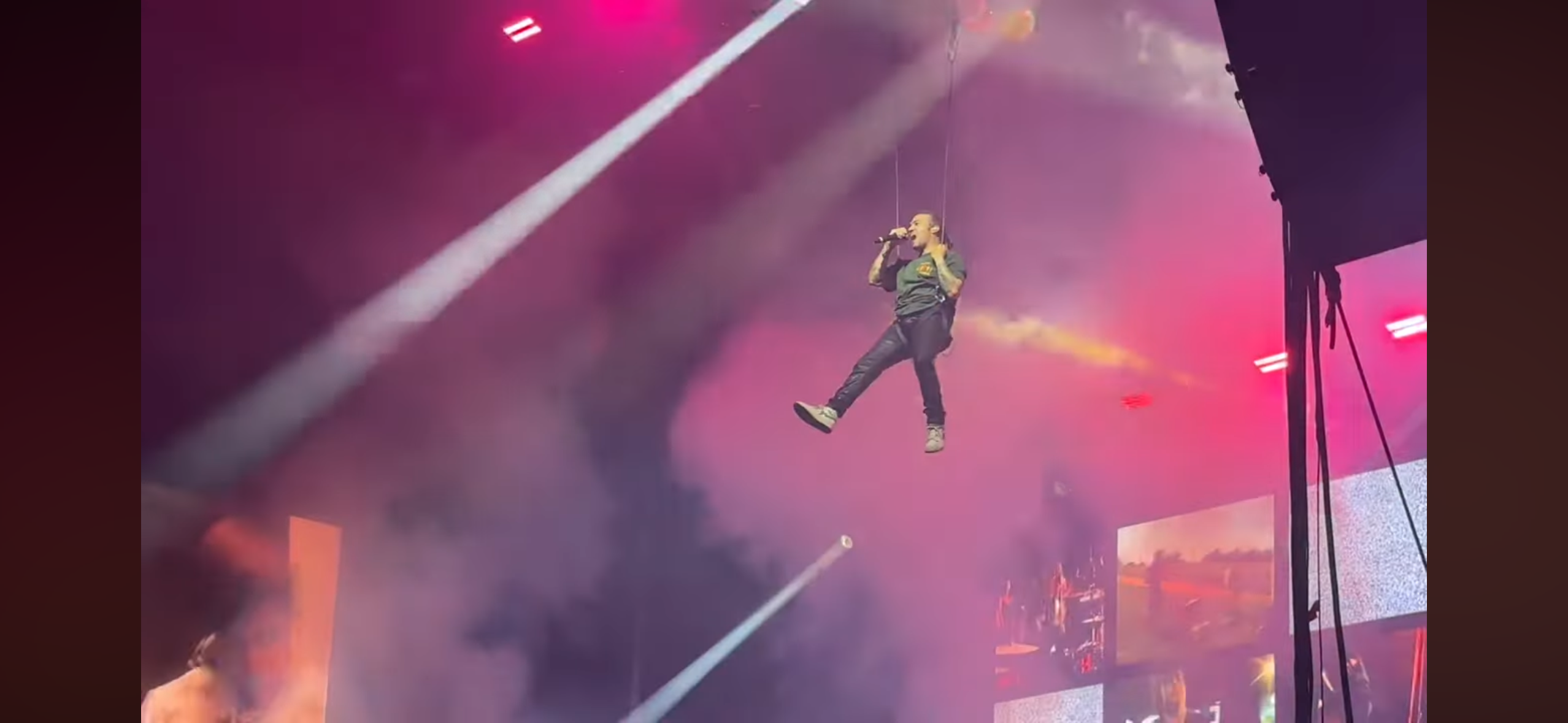 a musician is suspended in the air singing on stage during a concert. the stage is lit with spotlights, and the musician is surrounded by smoke. there is an audience in the background.