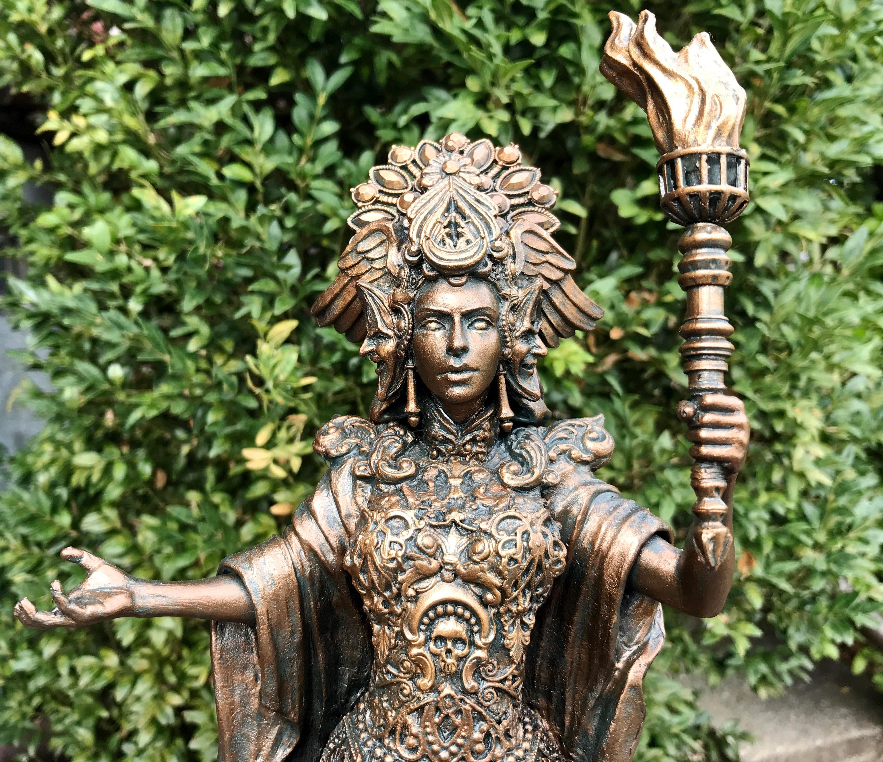 a bronzecolored statue of a woman in an ornate headdress and robes, holding a torch in her right hand and gesturing with her left. the statue is set against a backdrop of green foliage.