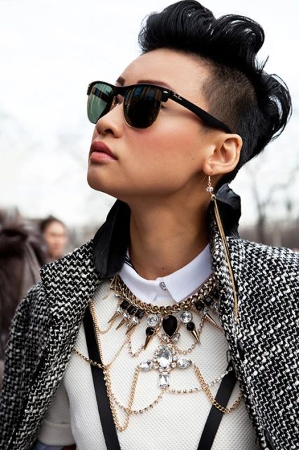 a woman with short black hair, styled in an undercut with the top styled upwards, wearing sunglasses, a white collared shirt under a white sweater, a large ornate necklace, long earrings, and a black and white patterned coat. she is looking up and to the left. the background is blurry and out of focus.