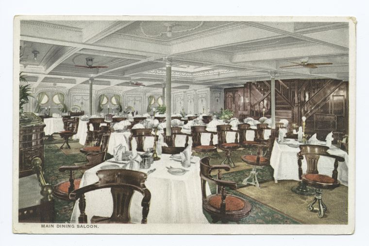 this image depicts the interior of a grand dining room, possibly on a ship, with tables set for meals, chairs, and architectural details such as columns, ceiling fans, and an ornate staircase.