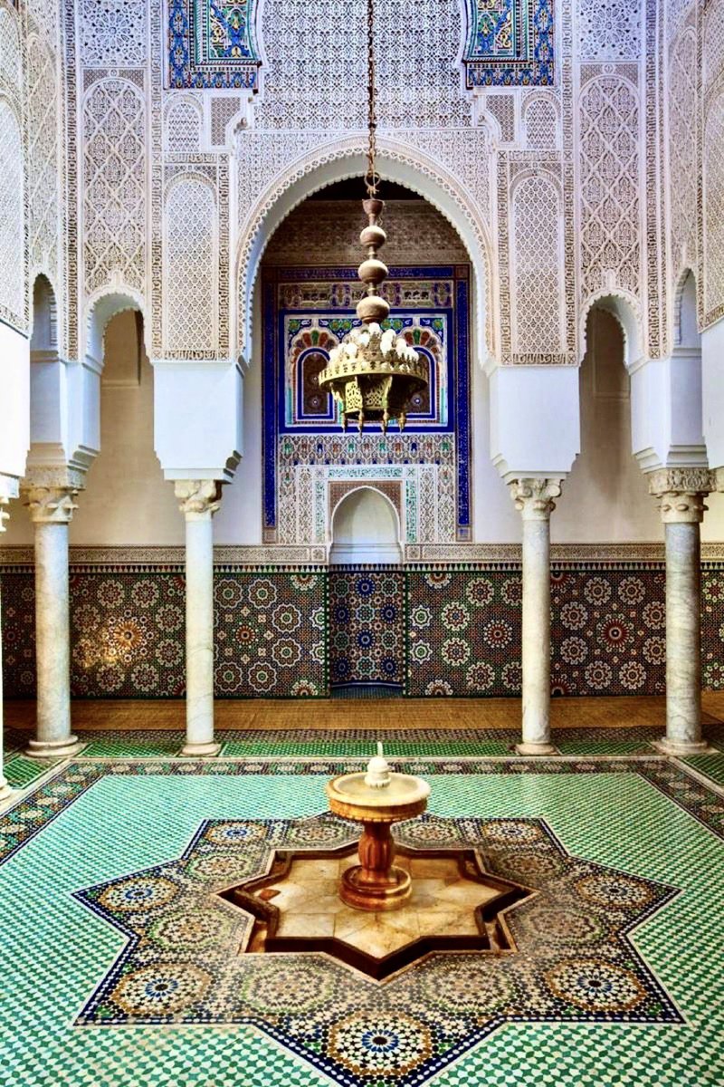 the image shows a beautiful islamic courtyard, likely in a mosque or similar building, featuring intricate mosaic tile work, decorative arches, and a central fountain. the walls are adorned with complex geometric patterns and ornate carvings, and a chandelier hangs from the ceiling. the overall design exemplifies moroccan or similar islamic architectural styles.