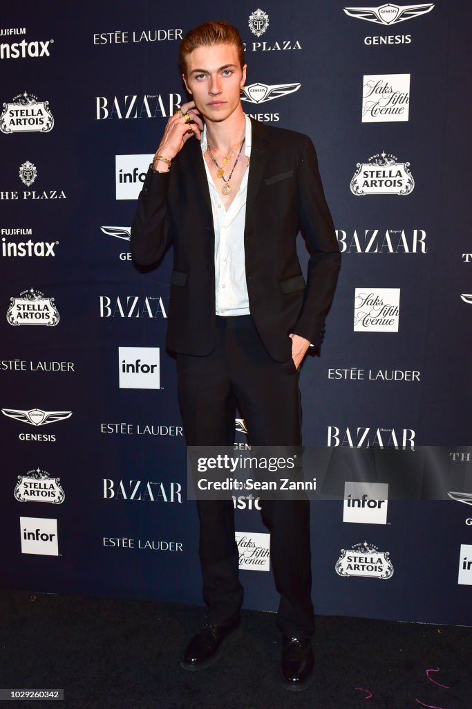 a man in a black suit stands in front of a backdrop featuring logos for brands like bazaar, genesis, stella artois, estee lauder, saks fifth avenue, and fujifilm instax. the man is wearing a necklace and dress shoes. he appears to be at an event or party.