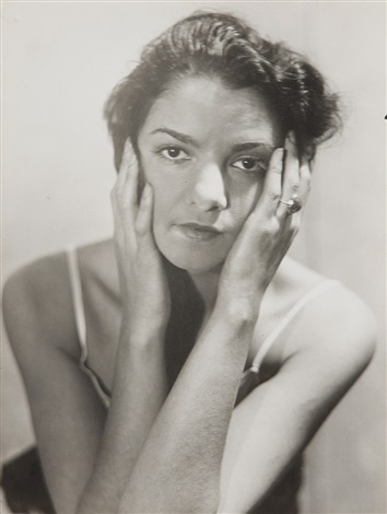 the image is a black and white closeup portrait of a woman.  she is positioned slightly angled, her hands gently cupping her face. her expression is serious and contemplative.  her dark hair is styled in a fashion typical of the mid20th century.  she appears to be wearing a simple, thinstrapped garment. the background is plain and unblemished, focusing all attention on the subject. the lighting is soft and subtle, highlighting the textures of her skin and hair.  the overall tone is elegant and classic.