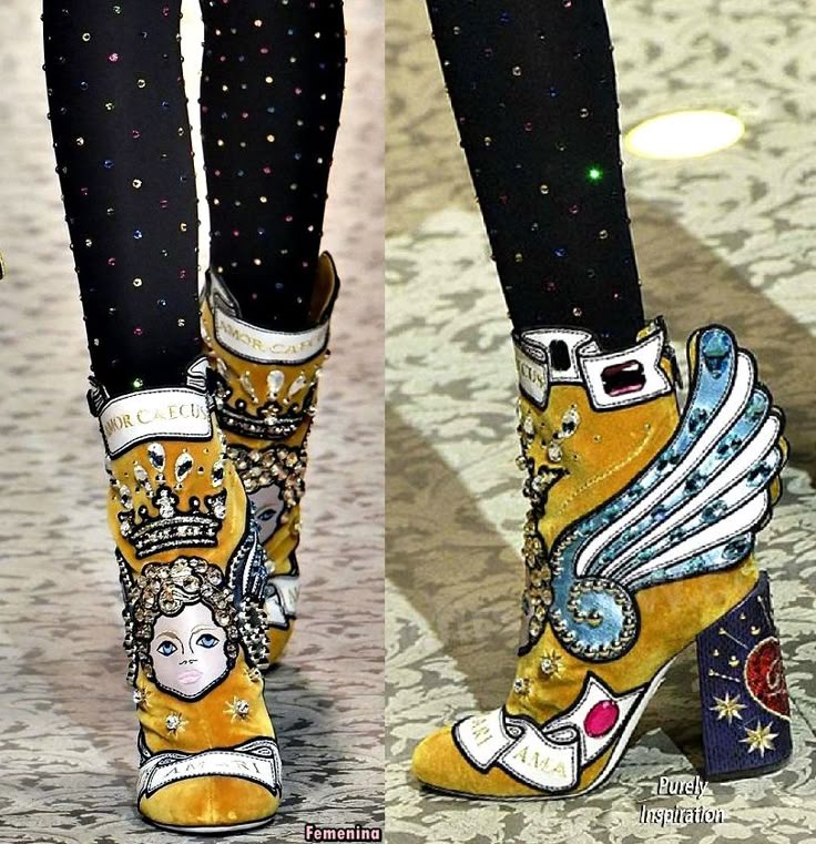 two pairs of extravagant yellow velvet ankle boots with elaborate designs, including a face with a crown, wings, rhinestones, and the words "amor caecus," are displayed on a patterned surface with legs wearing patterned tights.