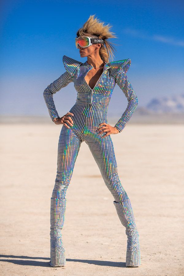 a woman is standing in a white desert wearing a silver jumpsuit, silver boots, and goggles.  she is striking a pose. the day is sunny, and the sky is blue. the woman is looking to the right.