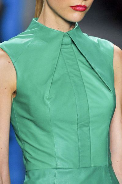 closeup view of a woman's torso, showcasing a mint green sleeveless leather dress. the dress has a distinct collared neckline with a tailored, structured design. the focus is on the texture and stitching of the leather. the model's makeup features bright red lipstick. the background is blurred, suggesting a runway or fashion shoot setting. the overall impression is one of high fashion and elegant style.