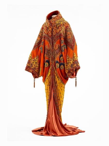 a fulllength photograph of a long, ornate dress on a mannequin. the dress is made of silk and satin in shades of orange, gold, yellow, and brown. it features intricate beading and embroidery throughout, creating a glamorous and flamboyant look. the dress is likely from the 1920s or 1930s, with an art deco influence. the dress is elegant and dramatic, making it suitable for a theatrical performance or a glamorous evening event. the dress is a testament to the craftsmanship and artistry of fashion designers from that era.
