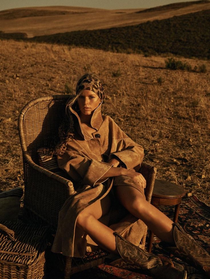 a fashion model sits in a wicker chair in a dry, grassy field at sunset. she is wearing a tan suede coat, cowboy boots, and a leopard print headscarf. the overall setting and attire create a stylish, relaxed, and warm atmosphere.  the image is professionally shot, likely for a fashion magazine or campaign. the warm tones of the sunset and earth colors are prominent.