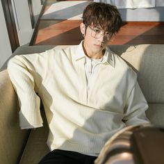 a young man with brown hair and glasses is sitting on a couch wearing a creamcolored, longsleeved pullover sweater with a collar. he is looking down. the image has a soft, natural light coming from a window. the background is a brown wooden wall.