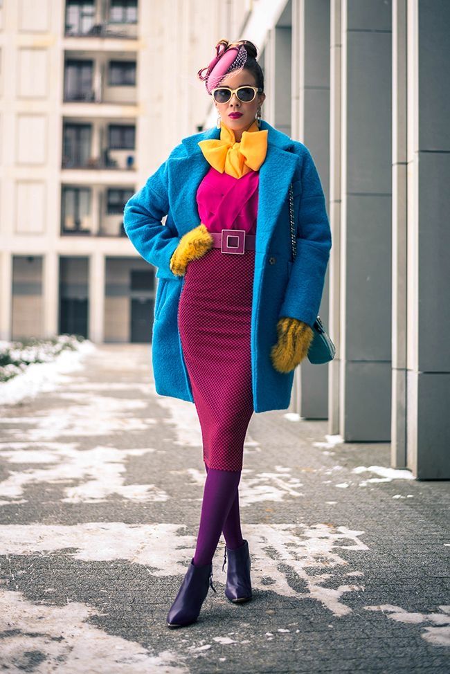 a woman stands outdoors in a colorful outfit featuring a bright blue coat, pink top and skirt, yellow accessories, purple tights, and boots. she is wearing a hat and sunglasses. the buildings are in the background. there is snow on the ground.