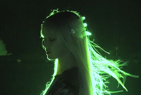 a woman with long white hair illuminated by glowing green neon light emanating from her hair. the background is completely black.