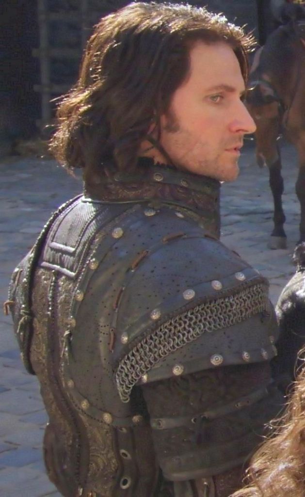 the image shows a man from the side. he has long brown hair and is wearing a black leather and metal armor suit. the armor appears to be from a historical period.