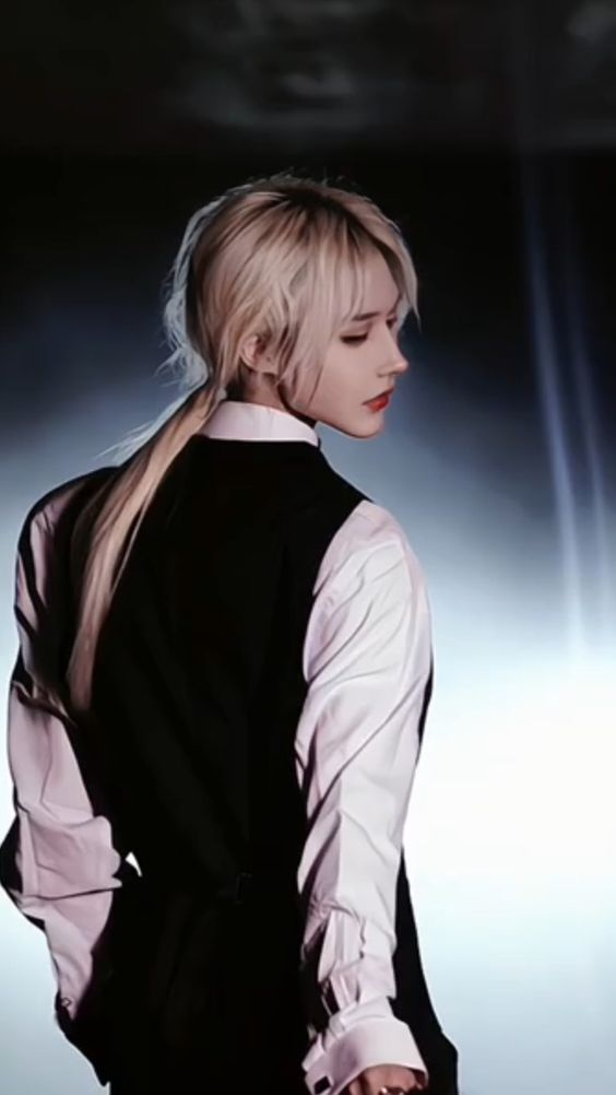 a person with long blonde hair pulled back in a ponytail, wearing a white shirt and a black vest is posing against a dark background in a studio setting. the model is facing right, looking away from the camera.