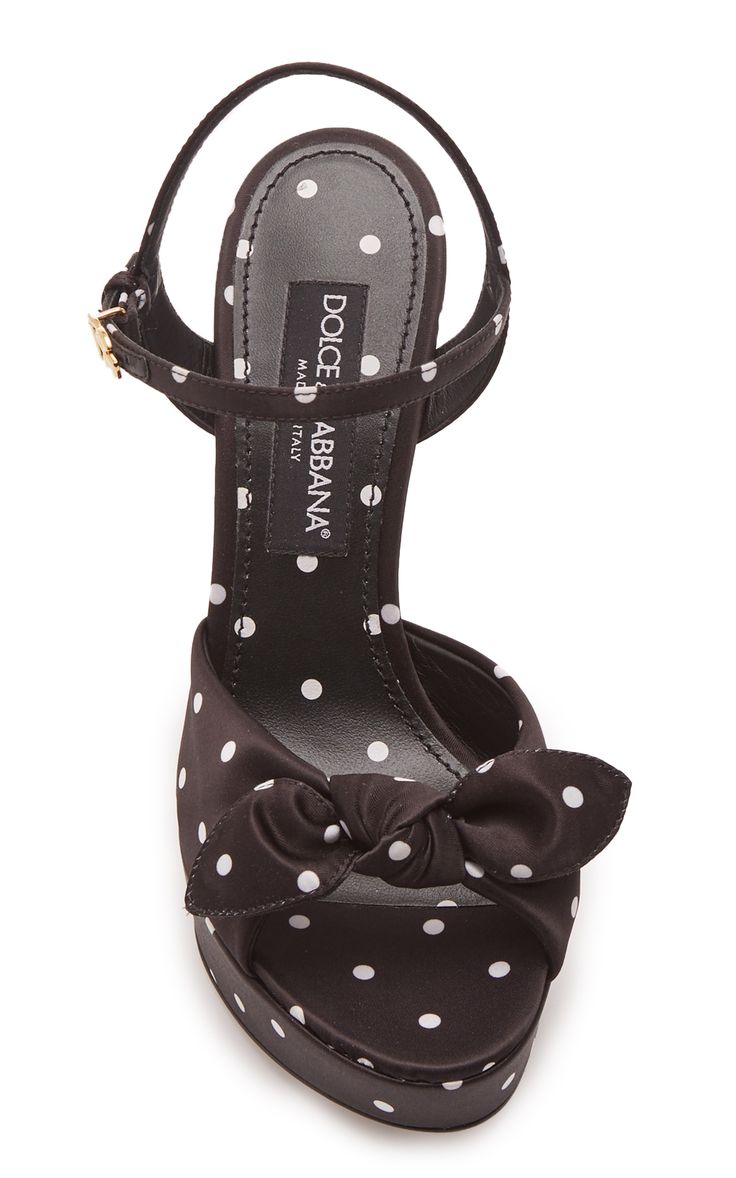 a studio shot of a pair of dolce & gabbana women's high heel sandals. the shoes are black satin with a white polka dot pattern.  they have a peep toe and a bow detail at the front.  the "dolce & gabbana" branding is visible on the insole.  the shoes are presented against a white background.
