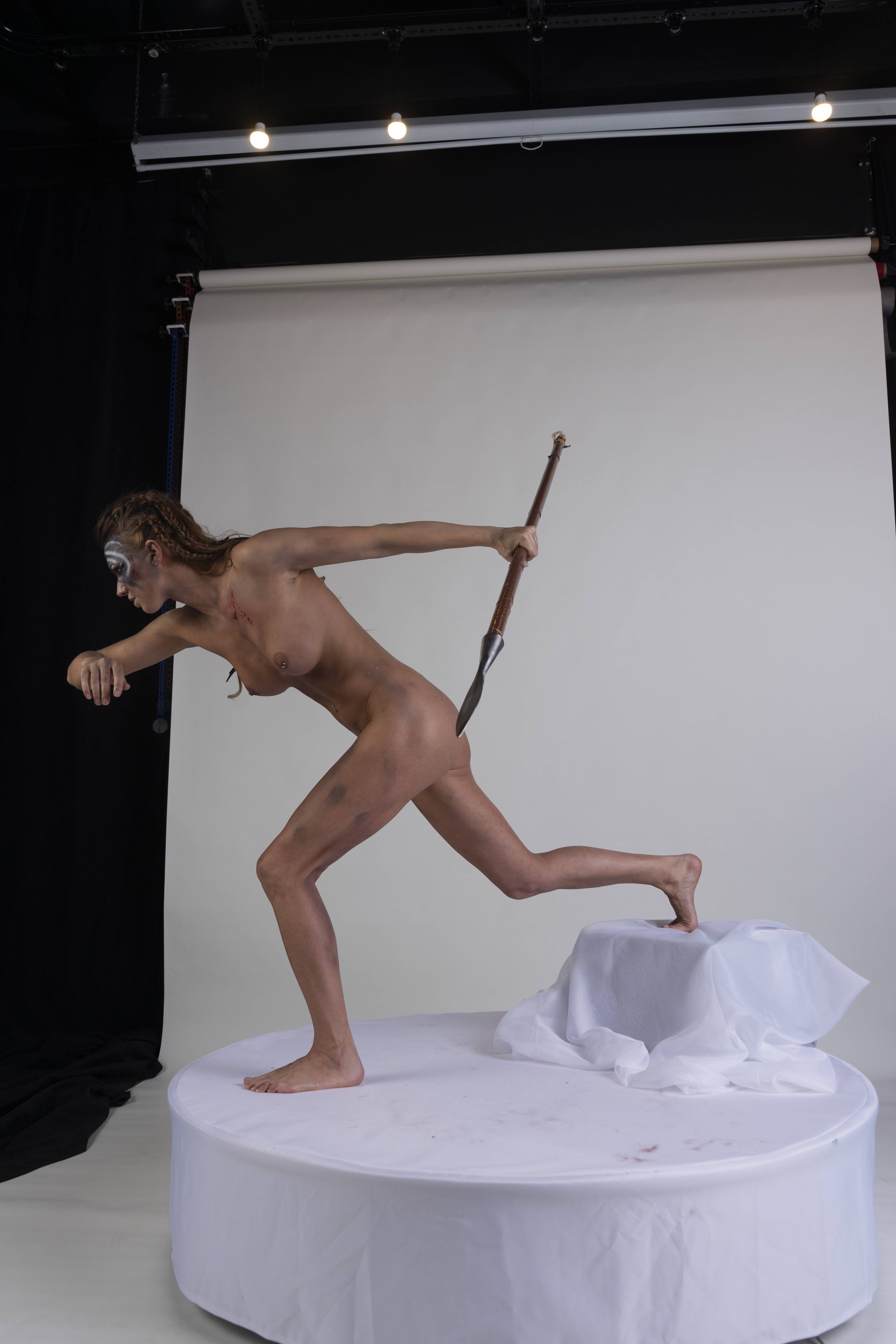 a nude female warrior with body paint and scars poses in a studio setting. she stands on a white platform holding a spear, with a white background behind her. her body is positioned in a dynamic pose, suggesting movement or action.