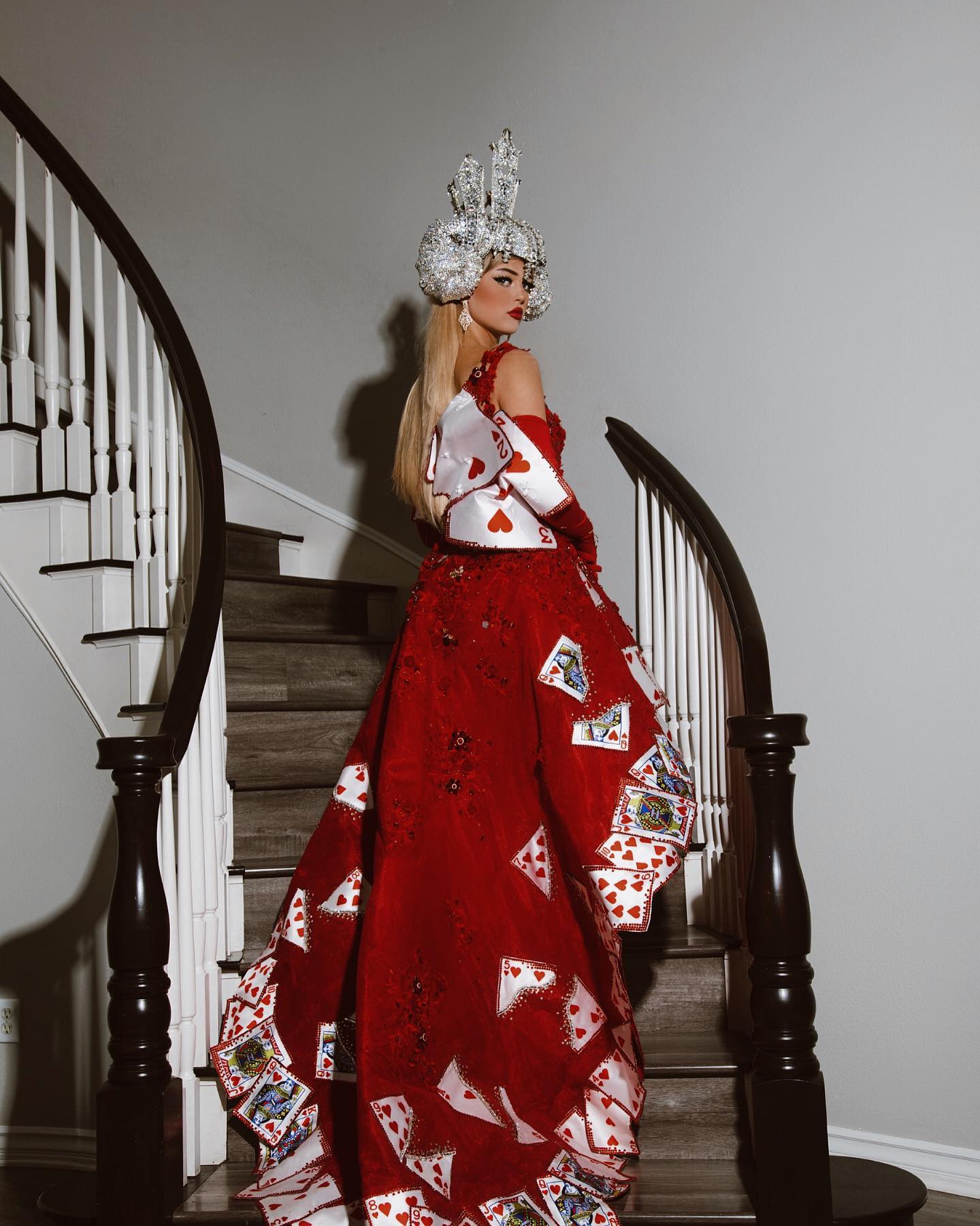 a woman dressed as the queen of hearts stands on a staircase, looking over her shoulder at the camera. she is wearing a red gown with playing cards attached to the skirt, and a silver headdress. the staircase has a white banister with a dark wood railing. the walls are a light gray.