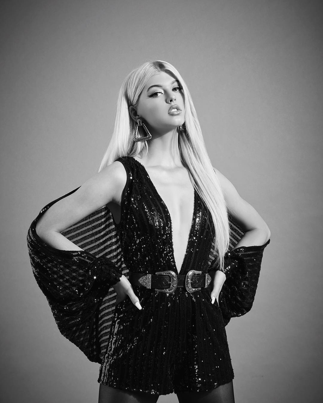 a black and white portrait of loren gray wearing a sequin jumpsuit with a deep vneck and a wide belt with two large buckles. she is also wearing triangular earrings. she is standing in front of a grey background with her hands on her hips.