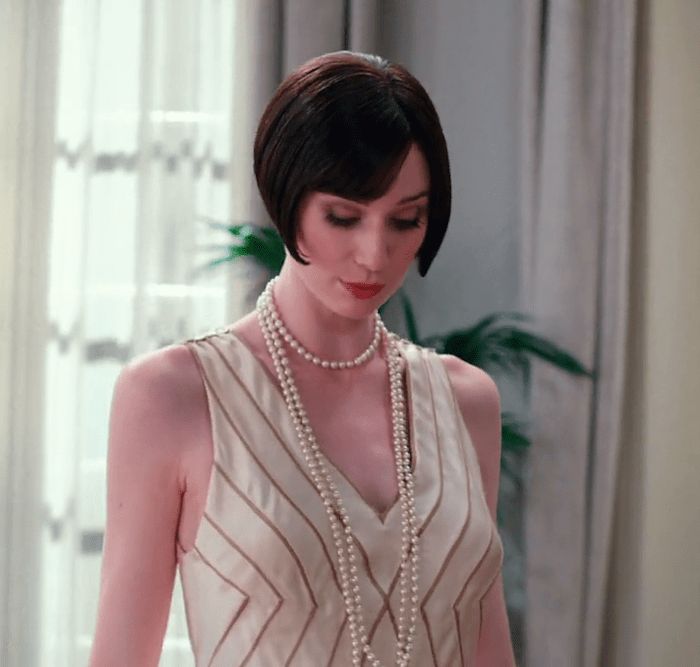 a young woman with a bob haircut, dressed in a white dress with a pearl necklace, stands indoors with a serious expression. she is looking down, her expression suggests a thoughtful or pensive mood. she is elegantly dressed in a style reminiscent of the 1920s. the background is light and blurry, focusing attention on the woman.