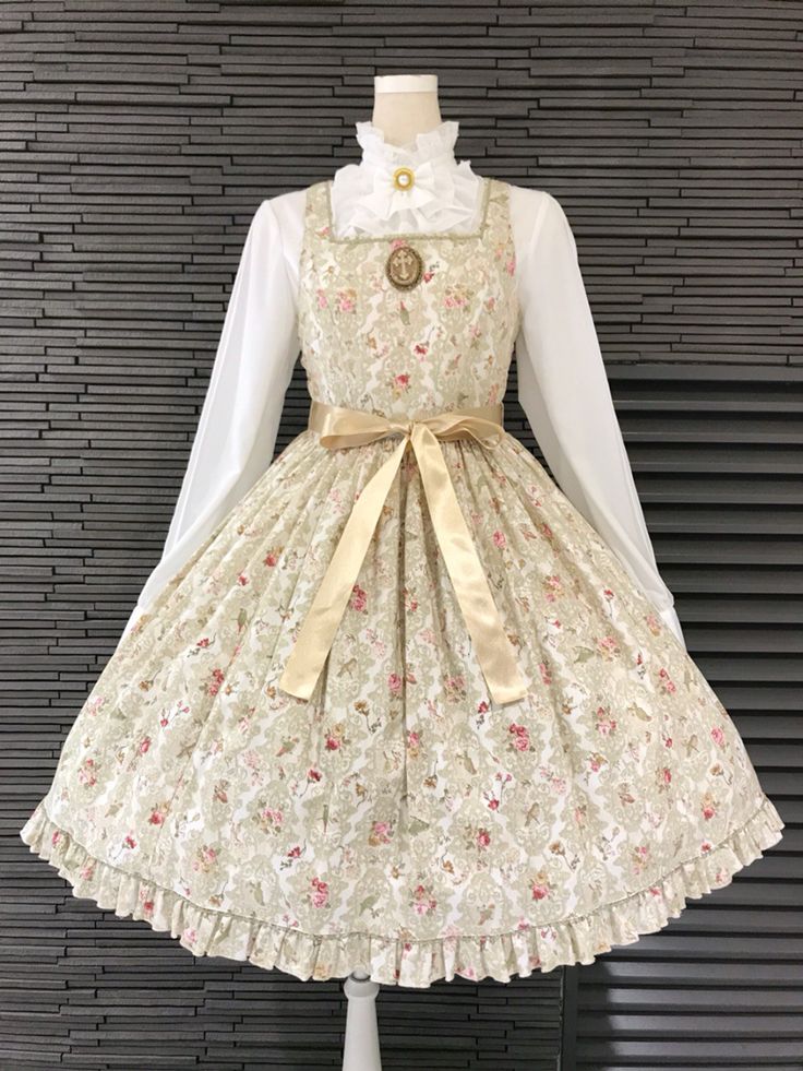 a full frontal view of a classic lolita fashion dress. the dress is displayed on a mannequin, showcasing a cream and pink floral print, a square neckline, delicate ruffles, and a matching satin ribbon tied at the waist. the mannequin is also wearing a white blouse with a high ruffled collar, complementing the overall vintage aesthetic.