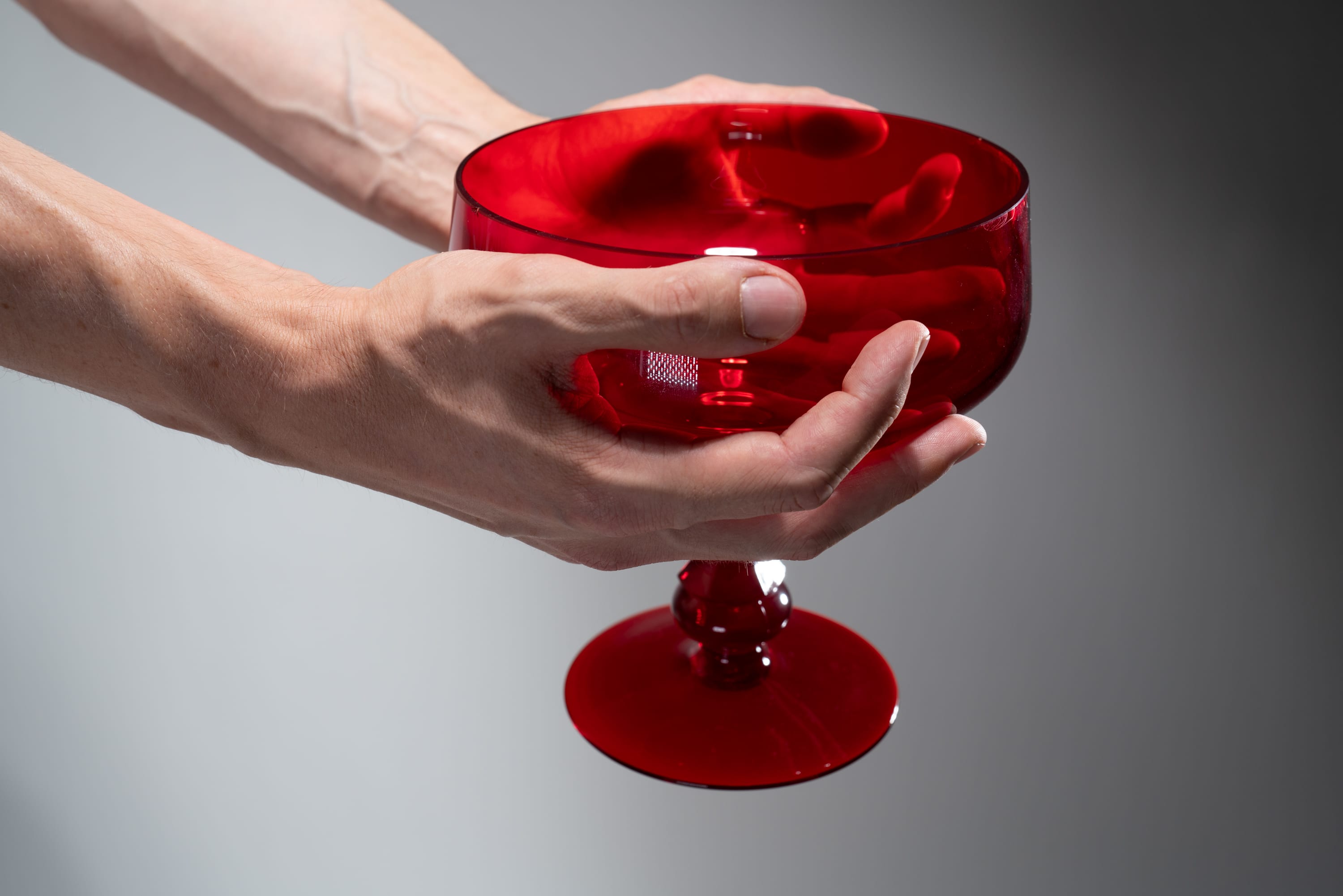 a pair of fairskinned hands gently holds a vibrant red glass bowl. the bowl is slightly tilted, offering a glimpse into its empty interior. the backdrop is a soft gray, allowing the rich color of the bowl to take center stage. the lighting is controlled and even, highlighting the smooth curves and reflective qualities of the glass.