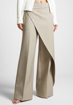 a woman is standing and wearing a beige pantsuit. the pants are wideleg and have a wraparound design. she is wearing a white cardigan over the pants and is not wearing a shirt underneath. she is standing in a studio setting.
