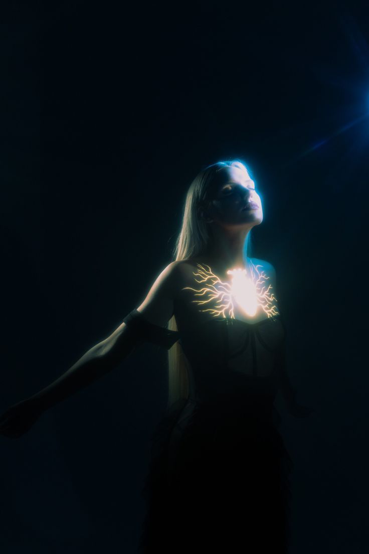 a woman with long blonde hair poses dramatically in a dimly lit space. she is wearing a dark outfit with a glowing heart design on her chest. the image has an ethereal and mystical quality to it.