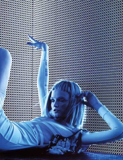 a woman in a futuristic pose in front of a blue grid wall, with her hair styled in a futuristic look. she is wearing a white outfit and has a cool, futuristic look. she is posing in a studio setting with blue and white light reflecting on her body and face.