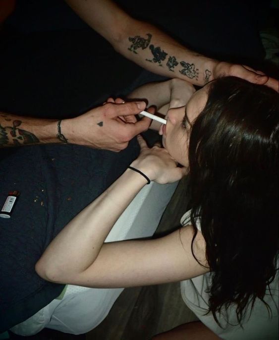 a woman with dark hair is taking a cigarette from a man's hand. the man has tattoos on both arms.