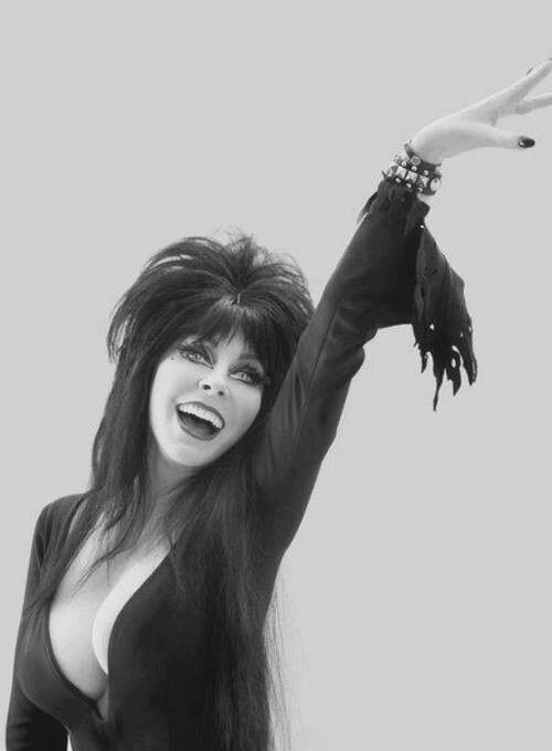 a black and white portrait of a glamorous woman in a black dress. she has long black hair, and she is smiling. she is posing with her arm raised, and she looks like she is about to strike a pose. she is dressed in a gothic style, with a black dress and long black hair.