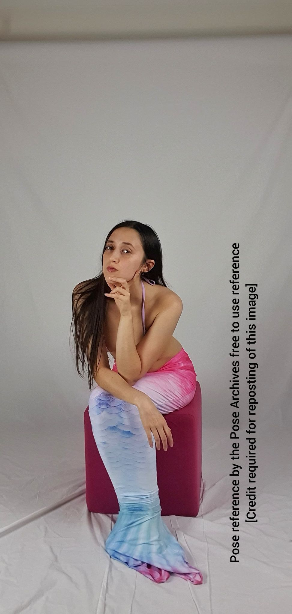 a young woman with long dark hair is posing in a studio setting. she is wearing a mermaid tail that is pink and blue. she is sitting on a pink cube and looking at the camera with a thoughtful expression. the background is a white backdrop and the lighting is soft.