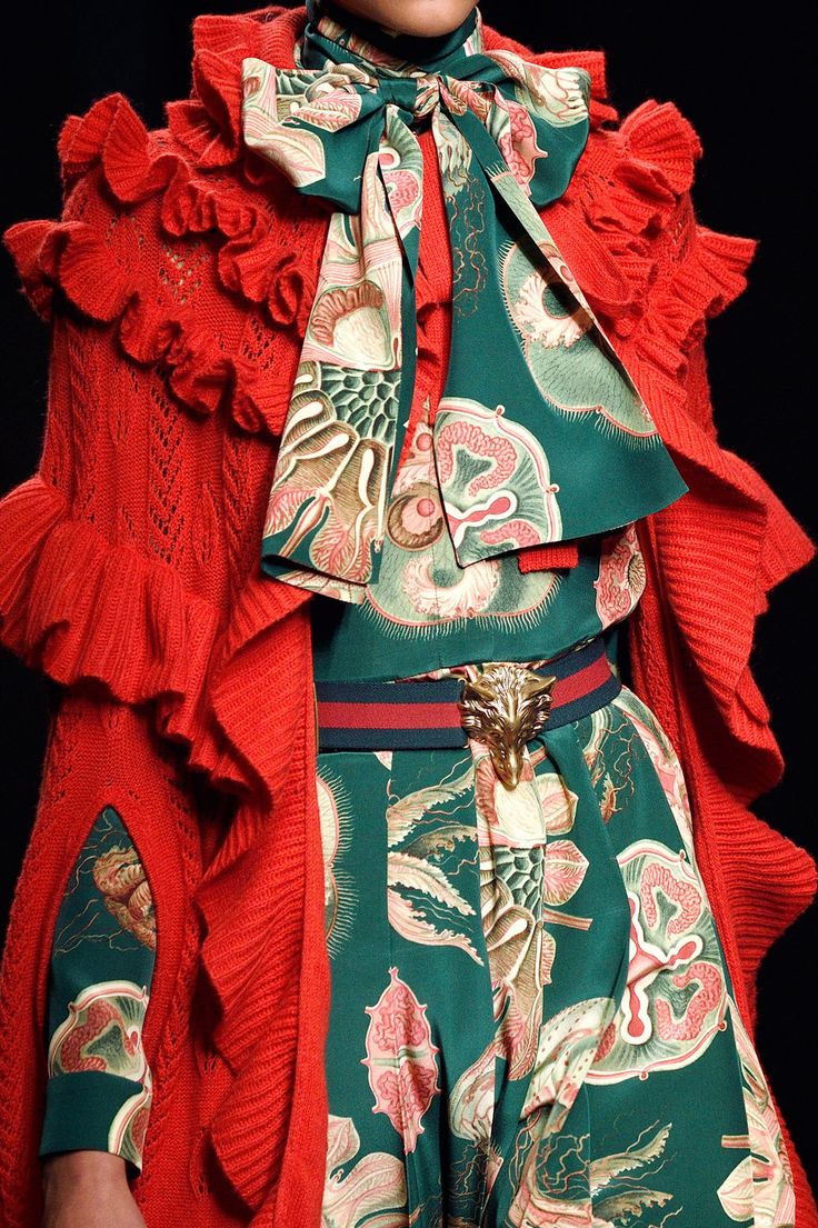 loseup detail shot of a model wearing a striking gucci outfit. the ensemble features a vibrant red ruffled knit cardigan over a teal silk dress adorned with a vintageinspired jellyfish and floral print. a green silk scarf with a similar print is tied in a bow at the neck. a distinctive gold fox head buckle accentuates the waistline. the image showcases the intricate details and rich textures of the fabrics. the overall aesthetic is high fashion and luxurious, typical of a gucci runway presentation.