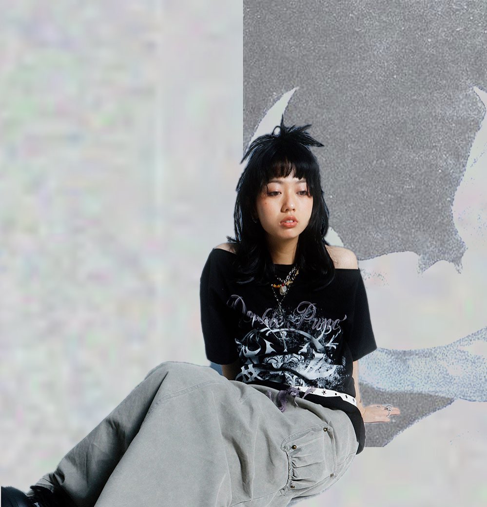 a young asian woman with dark hair is sitting on the floor against a textured gray wall. she is wearing a black offtheshoulder graphic tee, gray cargo pants, and a necklace. her pose is relaxed and casual. the image is a portraitstyle photoshoot.