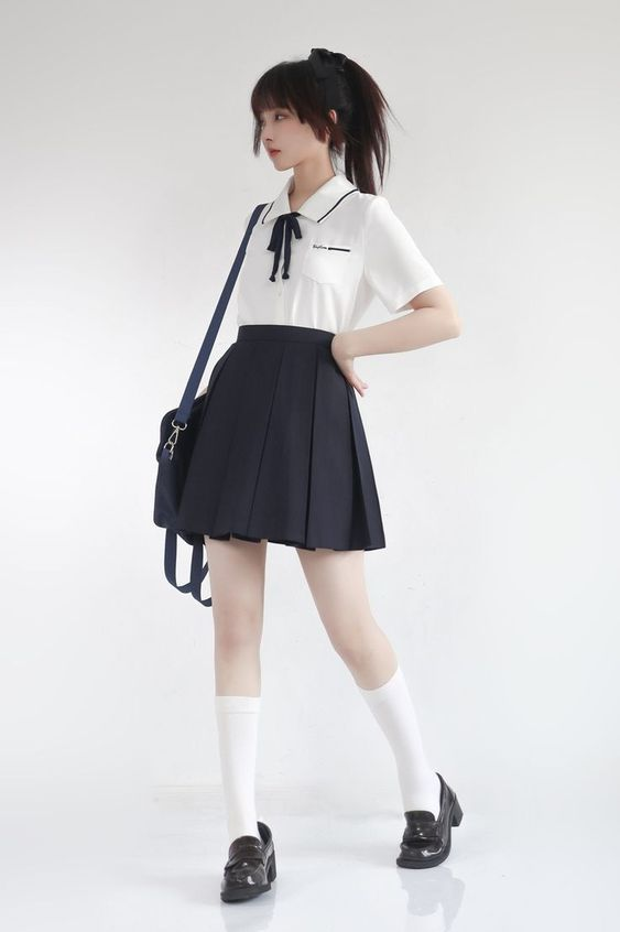 a young woman poses against a white background, showcasing a stylish school uniforminspired outfit. she wears a crisp white, shortsleeved blouse with a sailorstyle collar accented with a black ribbon. her navy blue pleated skirt falls just above the knee, complementing the classic silhouette. her accessories include black loafers, white ankle socks, and a black shoulder bag slung across her body. her hair is styled with a simple black bow, enhancing the youthful charm of the ensemble. the studio lighting illuminates her figure, highlighting the clean lines and tailored fit of the clothing.