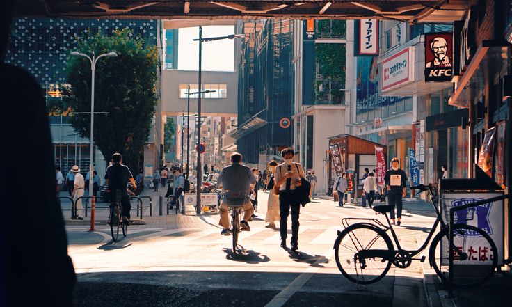 a bustling street scene in japan, bathed in bright summer sunlight. people are going about their day  some cycling, others walking. the street is lined with shops and buildings, and shadows are cast by the buildings and a streetlight. the photo is taken at eye level and captures the energy and movement of city life.