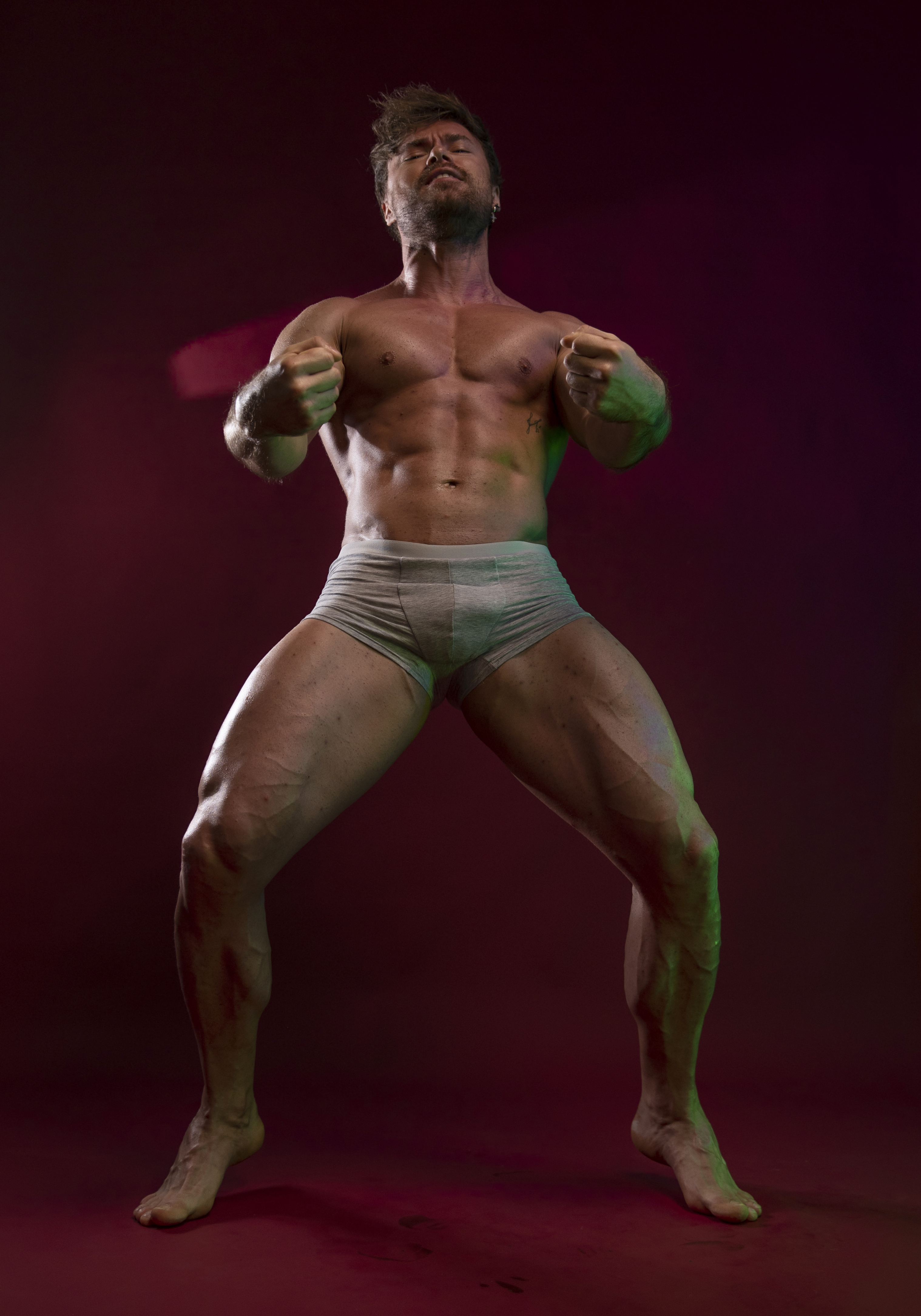 a muscular man in underwear is posing against a dark background. he is flexing his muscles and has a very athletic physique with visible veins. the lighting is dramatic, highlighting his physique.