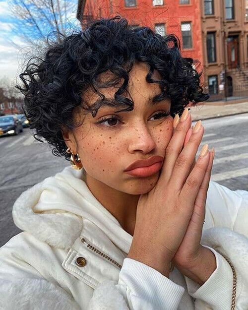 a closeup portrait of a woman with curly short hair and freckles. she is standing outdoors on a street, wearing a white jacket. her hands are clasped together in a praying or thoughtful pose, and she has a pensive expression on her face. the background shows an urban environment with buildings.