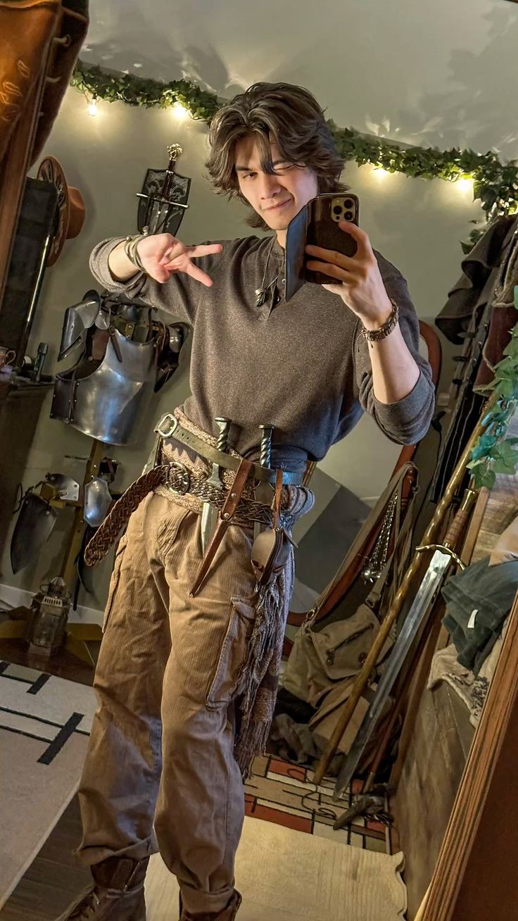 a person is taking a selfie. they are dressed in brown pants and a brown shirt and wearing a belt with swords and armor. they are holding up a peace sign.