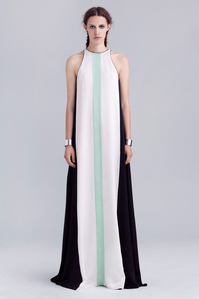 a fulllength studio shot depicts a slender woman wearing a long, sleeveless gown. the dress is primarily black on the sides with a white panel down the center, adorned with a thin, light green stripe vertically along the white section. the woman's wrists are accentuated with silver bracelets. her hair is styled in two braids, and she is facing forward against a plain, light grey background.
