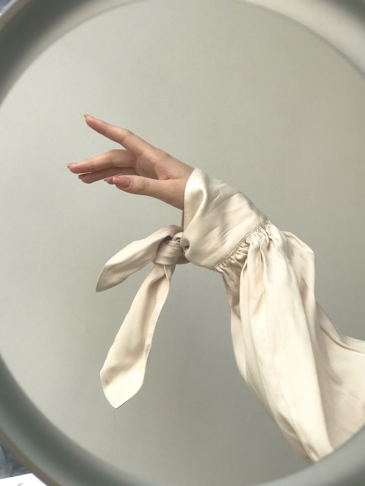 a closeup studio shot captures a hand gracefully poised within a neutral color palette. the subject's fingers are slightly spread, accentuating the soft, ivory tone of the skin. the wrist is adorned with a satin or silk sleeve, gathered at the wrist with a delicate tie or bow. the fabric has a subtle sheen, hinting at luxury and sophistication. the overall aesthetic is minimalist and fashionforward, emphasizing elegance through simplicity.