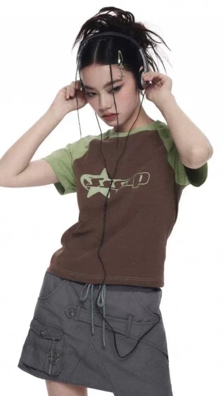 a young asian girl, possibly a teenager, is modeling a stylish outfit.  she's wearing brown and green raglansleeved tshirt with a graphic design and a grey cargostyle mini skirt. she has dark hair styled in a high ponytail and is wearing headphones around her neck. her expression is serious and focused. the background is plain white, making the outfit and the model the focal point of the image. the overall style is casual and slightly edgy, with a hint of 90s/y2k inspiration.