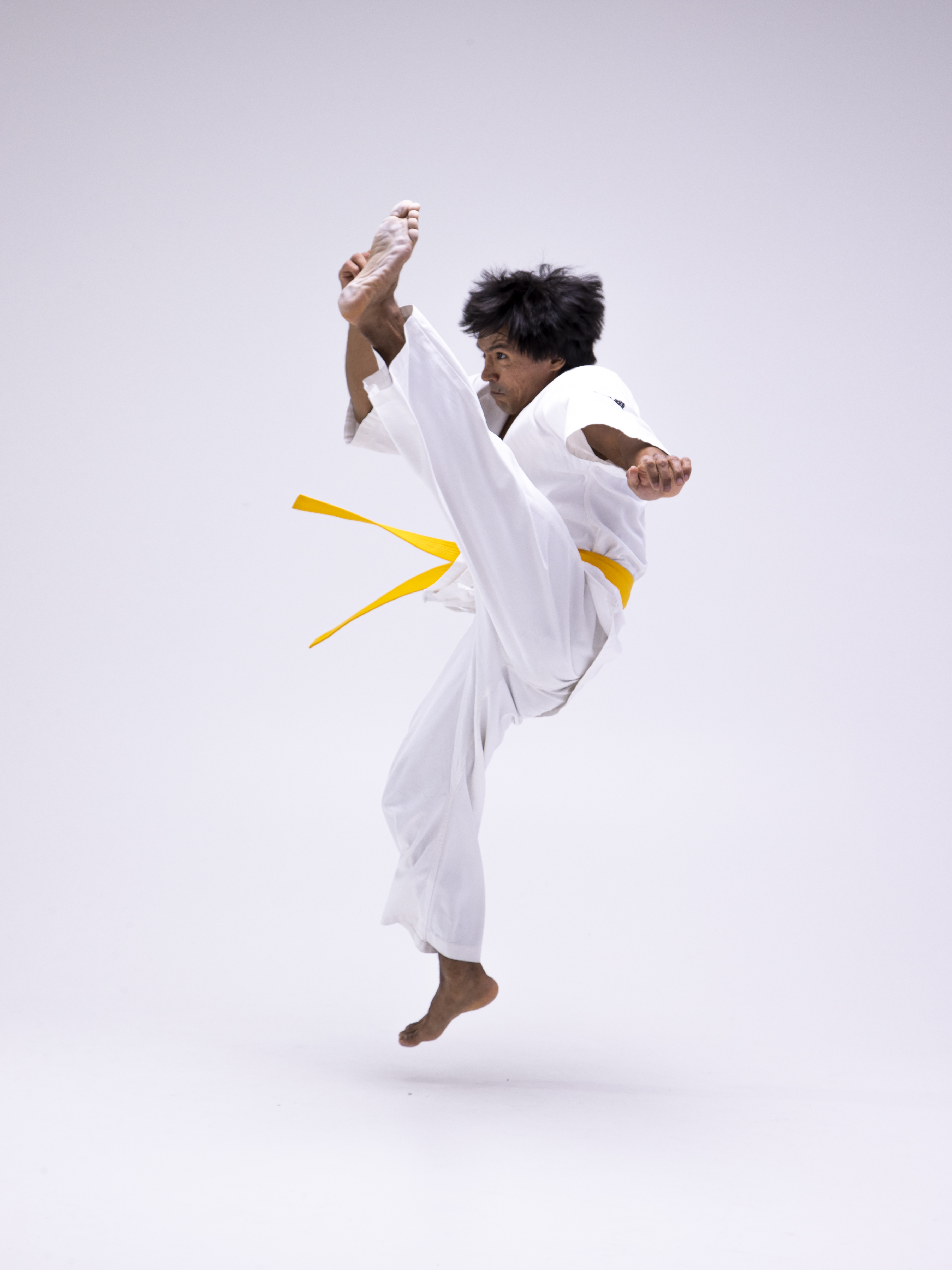 a man wearing a white karate gi and a yellow belt is captured in a dynamic studio shot, performing a high kick while airborne. he is barefoot, showcasing the action and movement inherent in the martial arts discipline of karate. the shot is a full shot, capturing his entire body against a white background.