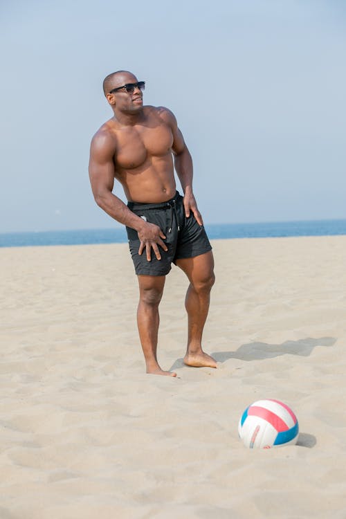 a muscular, shirtless black man in sunglasses and black shorts stands on a sandy beach, looking away from the camera. a volleyball sits in the sand near his feet.  the ocean is visible in the background. the scene suggests a sunny, summer day.